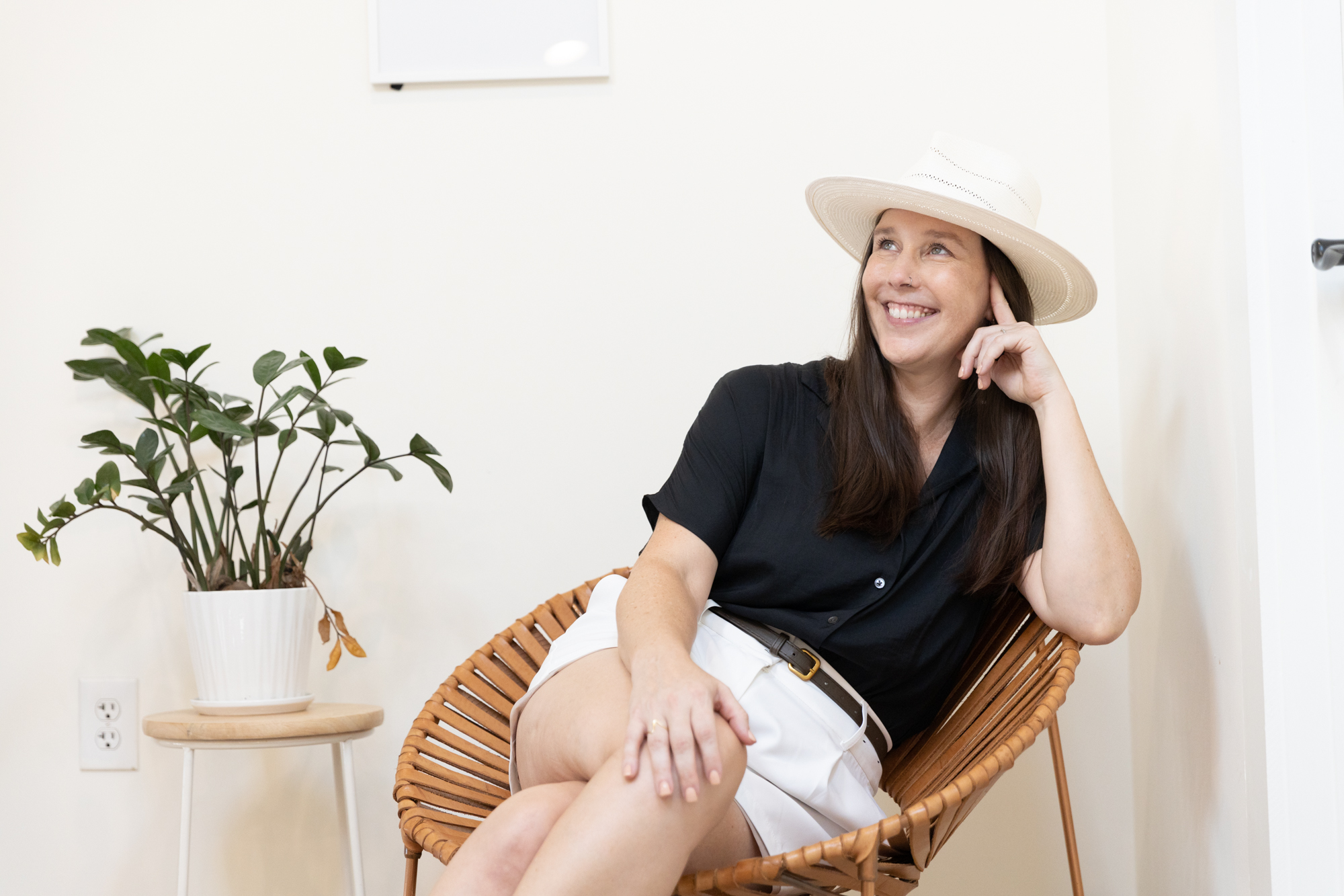 Branding Photographer sitting in a wicker chair next to plant wearing white straw hat, black blouse and white shorts
